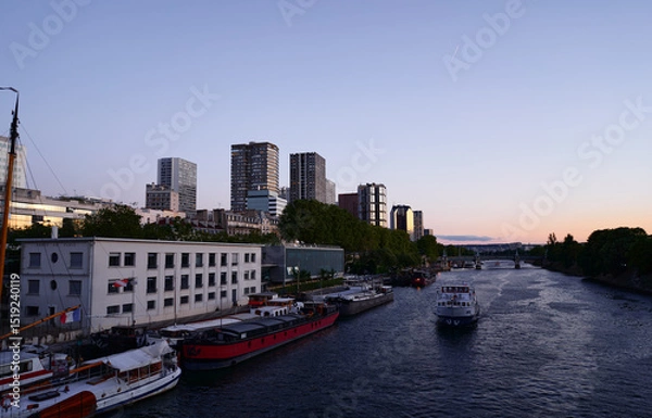 Obraz A peaceful riverfront scene showcasing boats alongside modern urban architecture, under a stunning sunset sky.