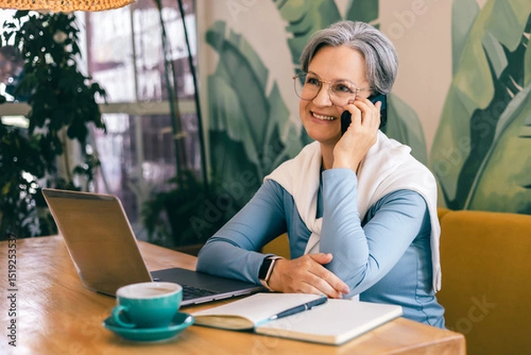 Fototapeta Confident mature woman smiling during phone conversation in modern workspace