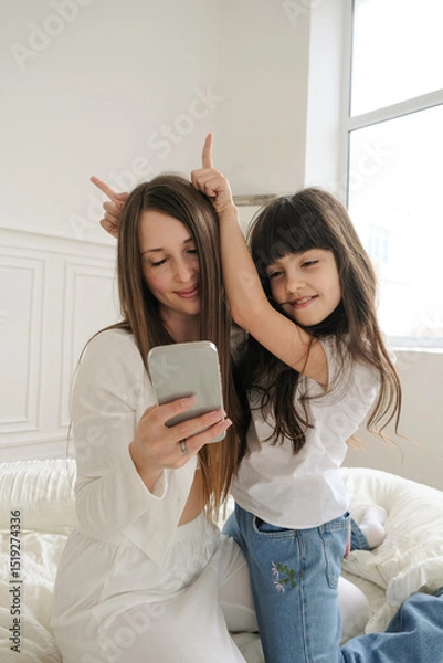 Fototapeta A mother and her young daughter sit close together, holding a smartphone as they chat with someone over video call. Their faces are lit with joy and affection, eyes focused on the screen. 