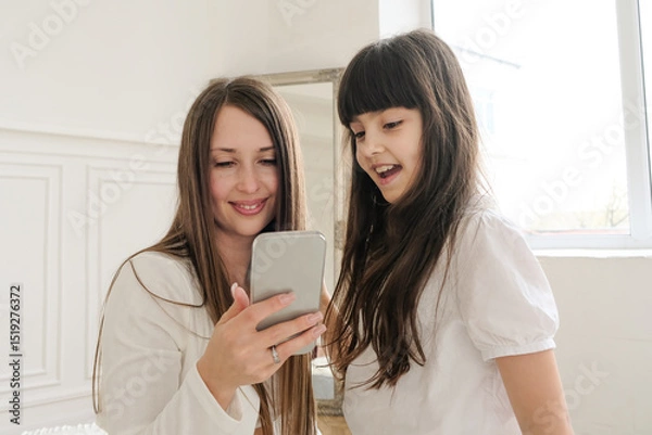 Fototapeta A mother and daughter are sitting together, looking at a phone while having a video call. They smile warmly, clearly enjoying the moment and the person on the other end of the screen. 
