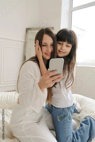 Fototapeta A mother and daughter are sitting together, looking at a phone while having a video call. They smile warmly, clearly enjoying the moment and the person on the other end of the screen. 