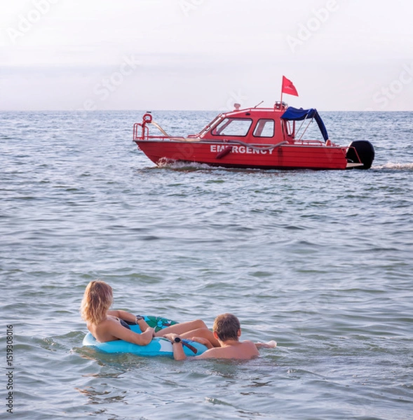 Fototapeta A young couple swimming in the sea with an inflatable circle watches as a red rescue boat passes by them