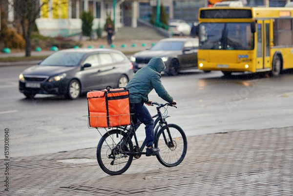 Fototapeta Fast food courier on bicycle riding through the streets in cold weather, timely delivery while navigating urban traffic. Delivery worker on bicycle transporting food orders. Man delivering pizza.
