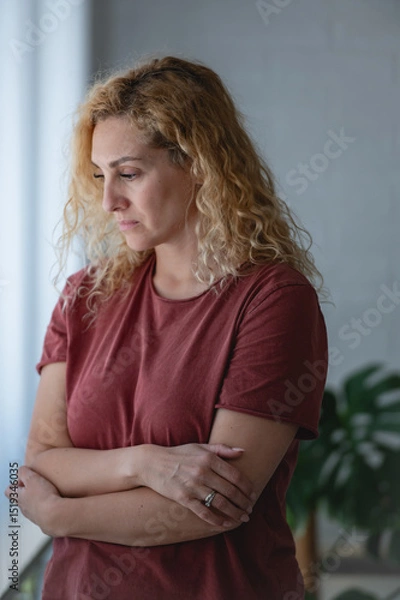 Fototapeta Thoughtful woman standing by a window with arms crossed, looking down with a serious expression, reflecting or feeling sad in a quiet home environment.