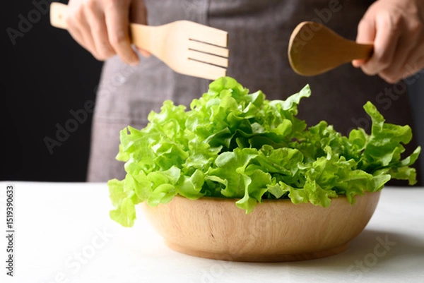 Obraz Organic green oak lettuce in wooden bowl with hand holding spoon and fork for cooking, Healthy eating