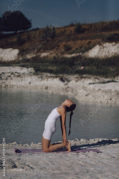 Obraz Woman in white sportswear doing camel pose on yoga mat by lake. Outdoor flexibility and wellness practice. Design for poster, banner, greeting card, invitation. Side view