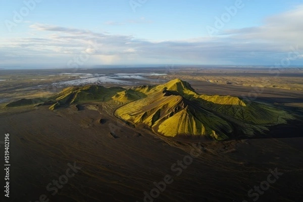 Fototapeta Aerial view of a volcanic mountain covered with green vegetation casting shadows on surrounding dark volcanic desert under a clear sky with scattered clouds