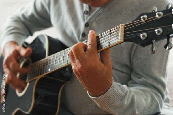 Fototapeta Senior man is playing guitar. Elderly male sitting at sofa and play music. Portrait of a gray haired mature 60s person with acoustic guitar, he is learning to play. Enjoying retirement life at home.