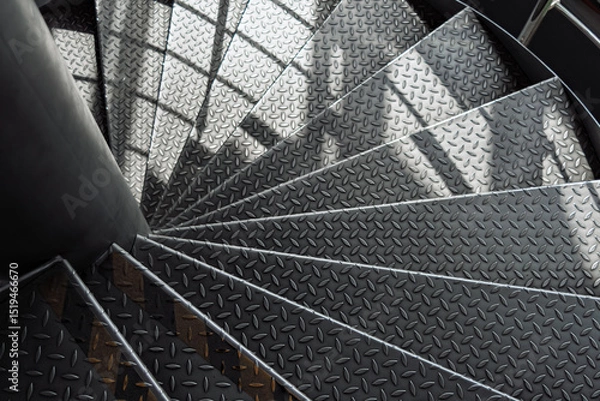 Fototapeta Curved metal staircase with textured diamond plate steps spirals downward, illuminated by soft daylight and dynamic shadows in a modern industrial interior.