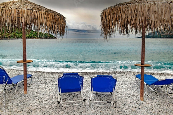 Fototapeta straw umbrella on a beach of white pebbles  with lounge chairs in front of a beautiful sea under cloudy sky in Peloponnese, Greece