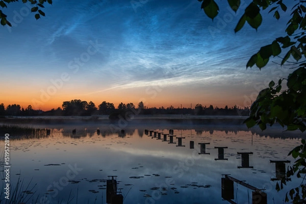 Fototapeta Tranquil lake at twilight with lily pads, pier remnants, framed by leafy trees, and glowing noctilucent clouds in a vibrant evening sky,slilver cloude