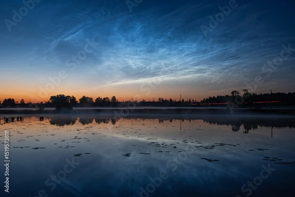 Fototapeta A calm lake at dusk with a misty pier, calm reflections, tree silhouettes and bright, noctilucent clouds in a colorful sky, silver clouds