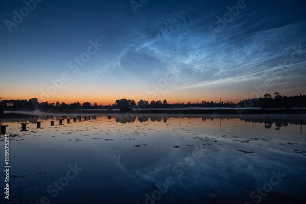 Fototapeta A calm lake at dusk with a misty pier, calm reflections, tree silhouettes and bright, noctilucent clouds in a colorful sky, silver clouds