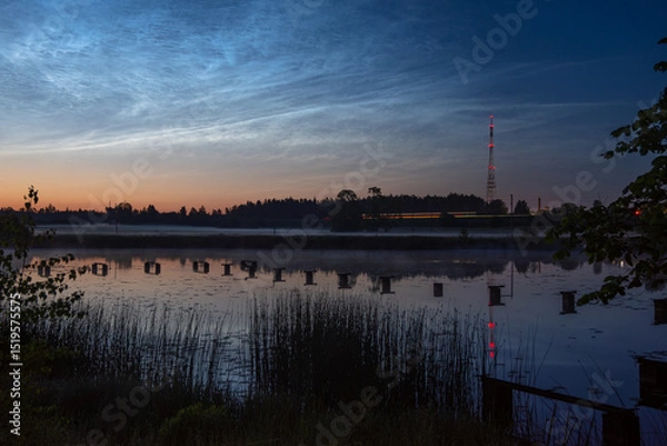 Fototapeta Twilight view of a calm lake with reeds, pier stubs, noctilucent clouds, and a red-lit radio tower reflected in the water near forest edge,silver clouds