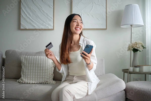 Obraz Casual asian woman sitting on sofa enjoying digital banking or mobile purchase on her smartphone.