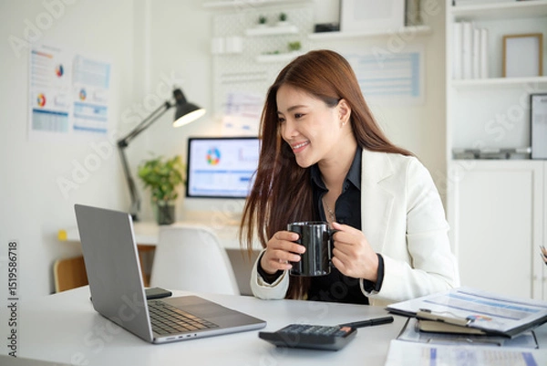 Obraz Businesswoman in white blazer holding a mug and smiling while looking at laptop in modern office setting.