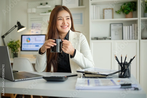Obraz Confident young businesswoman in modern office holding a black mug, relaxing and smiling while sitting at her desk surrounded by office supplies.