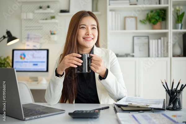 Obraz Smiling young Asian businesswoman holding a black coffee mug while taking a break in a bright modern office.
