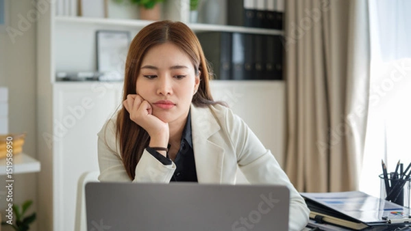 Obraz Young Asian businesswoman appearing fatigued and stressed while working on a laptop at the office desk.
