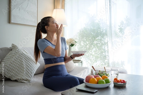 Obraz Young woman in sportswear relaxing on the sofa and enjoying a nutritious meal with a plate of fresh fruits and salad nearby.