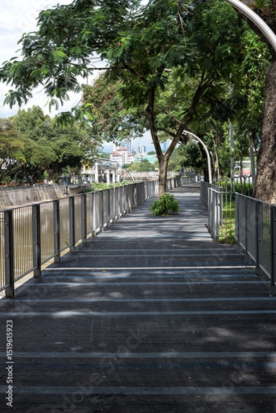 Fototapeta Scenic Riverside Walkway in Brickfields, Kuala Lumpur. The pathway runs parallel to the river, with tall buildings and tropical trees in the background.