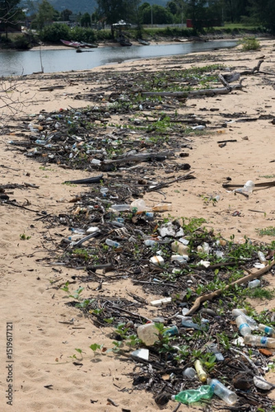 Fototapeta Polluted Beach with Plastic Waste and Debris. A sandy beach covered with scattered plastic bottles, waste, wood debris, and vegetation.