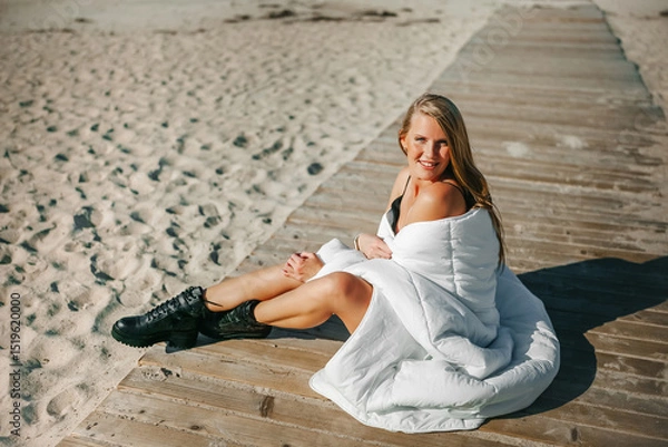 Fototapeta A serene beach moment with a woman wrapped in white blanket enjoying the sun on a wooden dock by the sea at midday