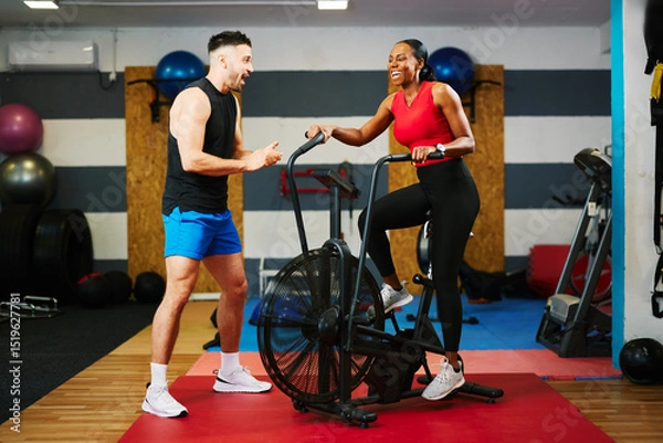 Obraz Woman using air bike with personal trainer assisting her during training session in gym