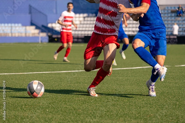 Fototapeta Two soccer players in red and blue uniforms competing for the ball during a match on a green artificial turf field.