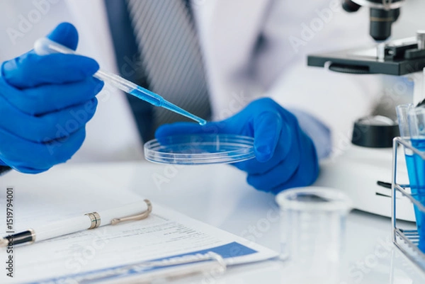 Obraz Scientist analyzing samples with pipette in laboratory. Scientist wearing blue gloves is holding a pipette with blue liquid over a petri dish, conducting research in a laboratory setting.