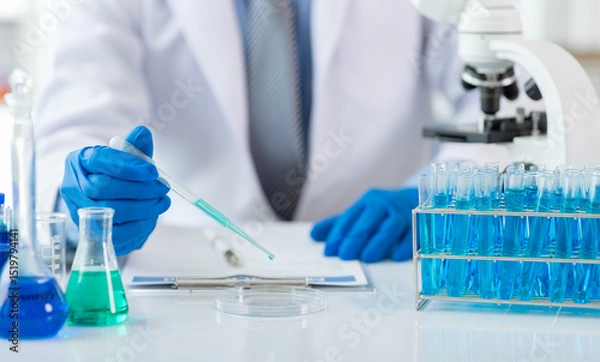 Obraz Scientist analyzing samples with pipette in laboratory. Scientist wearing blue gloves is holding a pipette with blue liquid over a petri dish, conducting research in a laboratory setting.