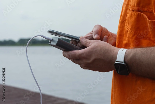 Obraz Man charging smartphone with power bank by lake using USB cable in summer outdoor. Concept modern lifestyle, mobile technology, travel, and connectivity on the go.