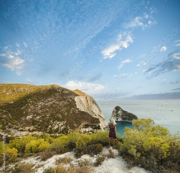 Obraz A traveler stands on a scenic cliff in Zakynthos, gazing at the azure waters and dramatic landscape. The vibrant summer sky enhances the serene atmosphere and natural beauty