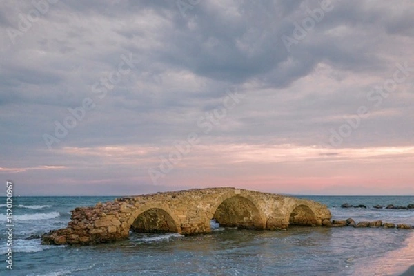 Obraz At the edge of the azure sea, an ancient stone bridge emerges from the waves as dusk casts warm hues across the horizon. Zakynthos summer tourism reveals captivating coastal charm