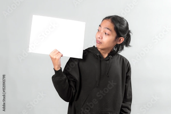 Fototapeta Asian man in black hoodie holding a blank white sign while looking at it with a curious expression, isolated on gray background, perfect for advertisement or message mockup.
