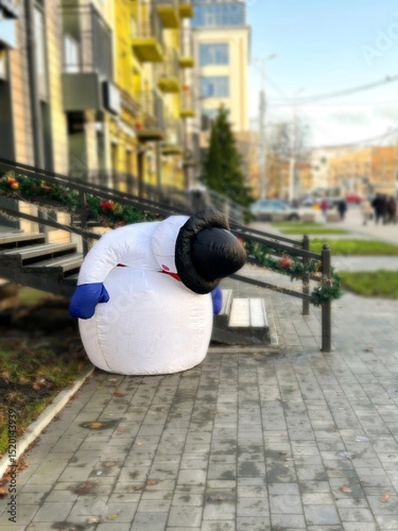 Fototapeta Broken life-sized snowman mascot decorates city center during Christmas season. Image with humor. Broken snowman illustrating poor Christmas and winter without snow.