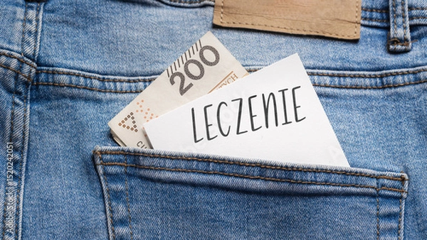 Obraz White card with a handwritten inscription "Leczenie", put into the pocket of blue pants jeasnow, next to Polish banknotes PLN (selective focus), translation: Treatment
