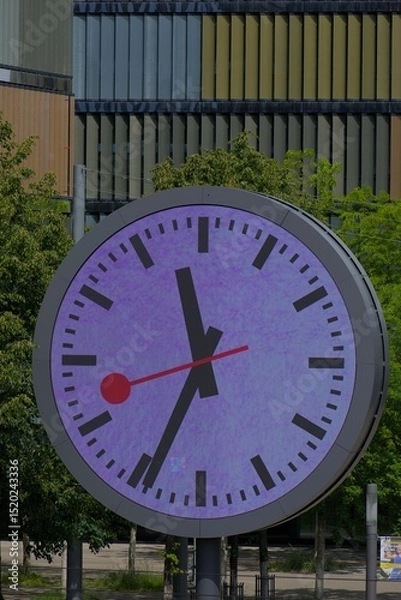 Fototapeta Iconic round clock face, vibrant purple hues, and classic black hands against lush greenery. A symbol of precision and timeless design in an urban setting.