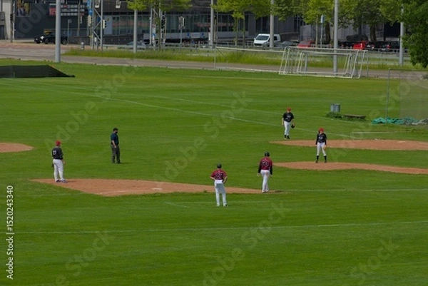 Fototapeta An expansive green field hosts a baseball game, capturing the sport's anticipation and strategic play under a bright, open sky.