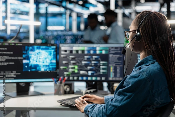 Fototapeta Data center support expert talking in headset microphone, supporting engineering team doing equipment maintenance. Employee in server farm coordinating technicians using mic