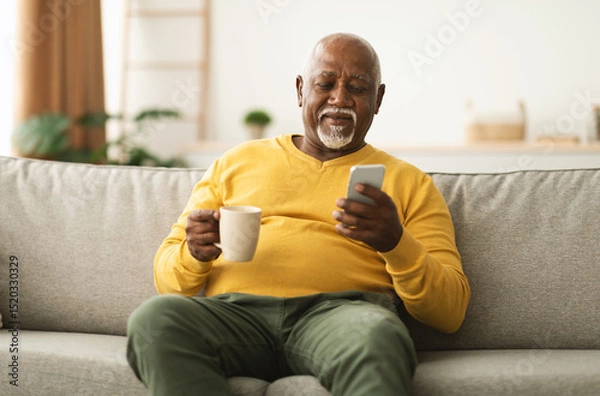 Fototapeta Mature African American Male Using Cellphone Browsing Internet And Texting Communicating Online In Social Media App Sitting On Sofa At Home. Senior Man Drinking Coffee And Scrolling News On Phone