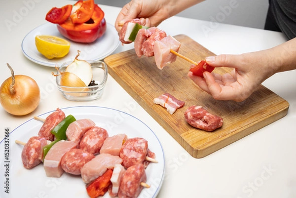 Fototapeta Hands of female chef preparing meat and vegetable skewers with pork, bacon, red and green bell peppers, and onions on a wooden cutting board in kitchen