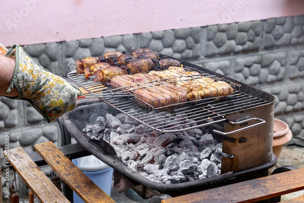 Fototapeta Close up hand of chef wearing a heat resistant glove skillfully adjusting meats and potatoes on a charcoal barbecue grill, creating a delicious outdoor meal for family and friends