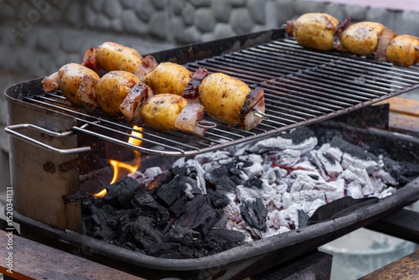 Fototapeta Deliciously grilling potatoes and bacon skewers over hot coals on a barbecue grill, creating a mouthwatering summer meal filled with smoky flavors and charred goodness