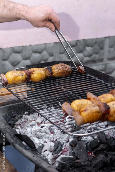 Fototapeta Hands of chef grilling skewers of potatoes and bacon over hot coals on an outdoor barbecue grill, creating a smoky and flavorful meal perfect for summer picnics and gatherings