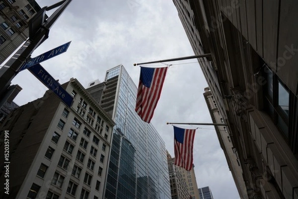 Fototapeta View looking up at tall city buildings with American flags flying on flagpoles against a cloudy sky at the intersection of Fifth Avenue