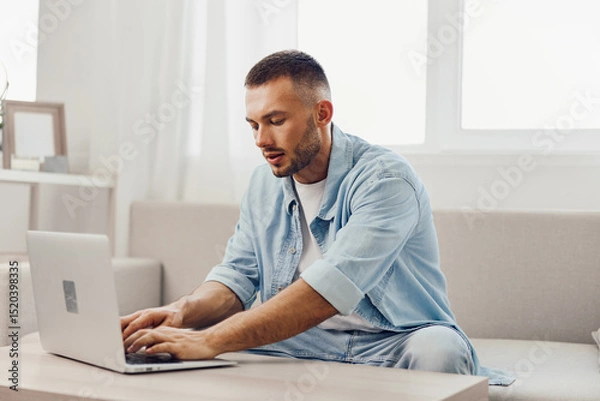 Fototapeta Focused man working on laptop with casual attire in a bright, modern living room, conveying a sense of productivity and creativity in a home office setting.