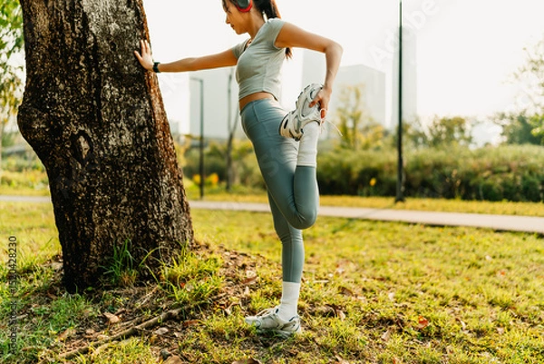 Fototapeta A woman dressed in workout clothes is wearing headphones and listening to music while stretching her muscles before exercising, before running, or after exercising.