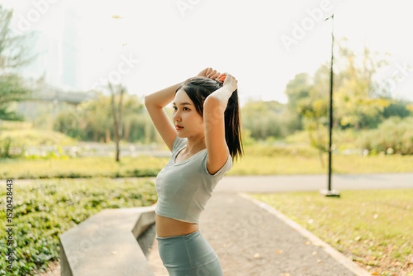 Fototapeta A woman in sportswear stands in a park, stretching with arms raised, enjoying a sunny day outdoors and preparing for a workout, surrounded by greenery and natural light.