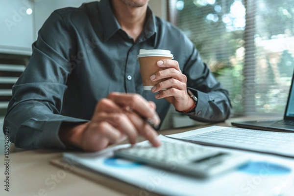 Fototapeta Businessman drink hot coffee. An accountant is seated at a desk in the office, utilizing a calculator to determine balances, profits, currency conversions, and costs.